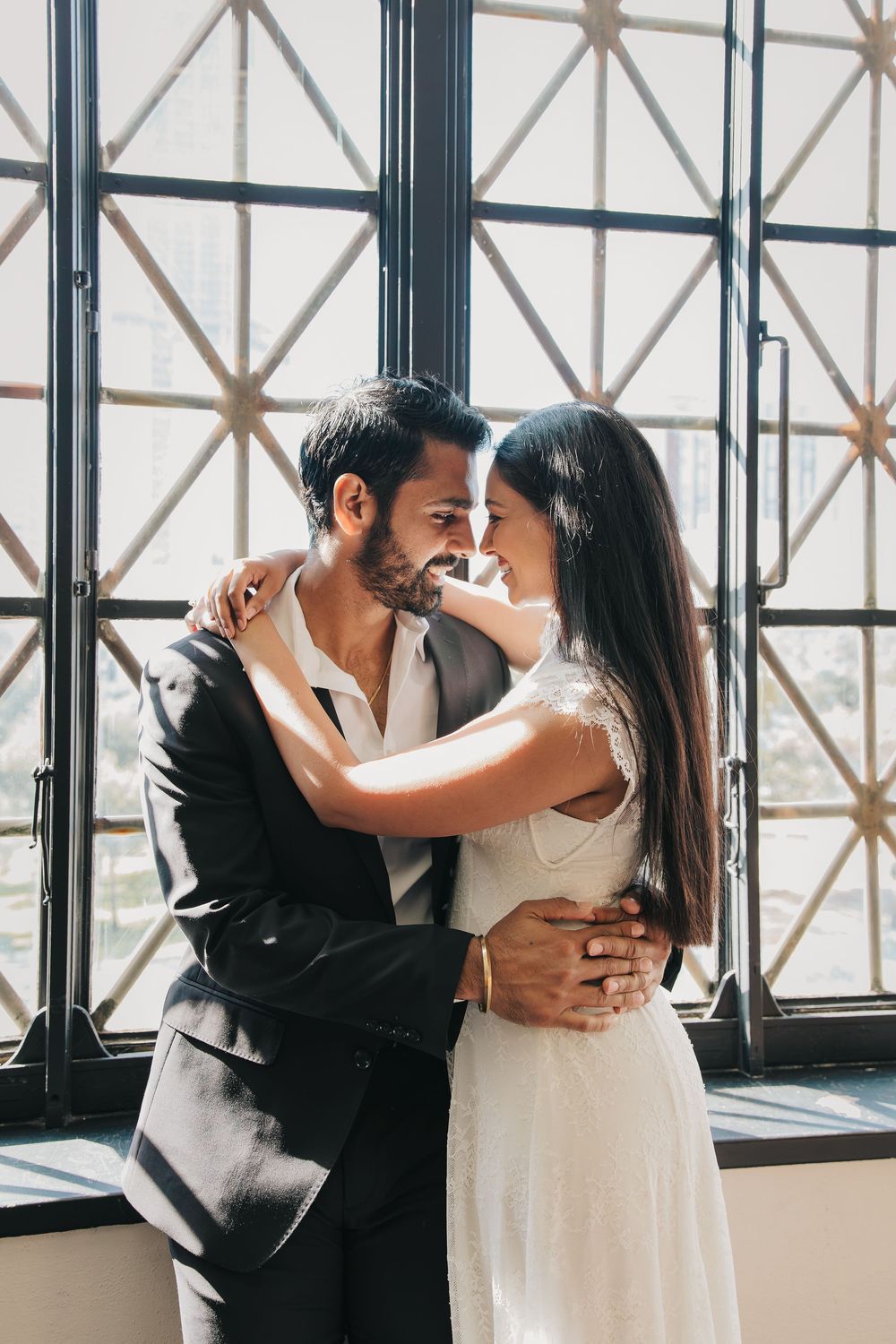 A romantic couple in wedding attire shares intimate moments in front of industrial windows in an urban setting.