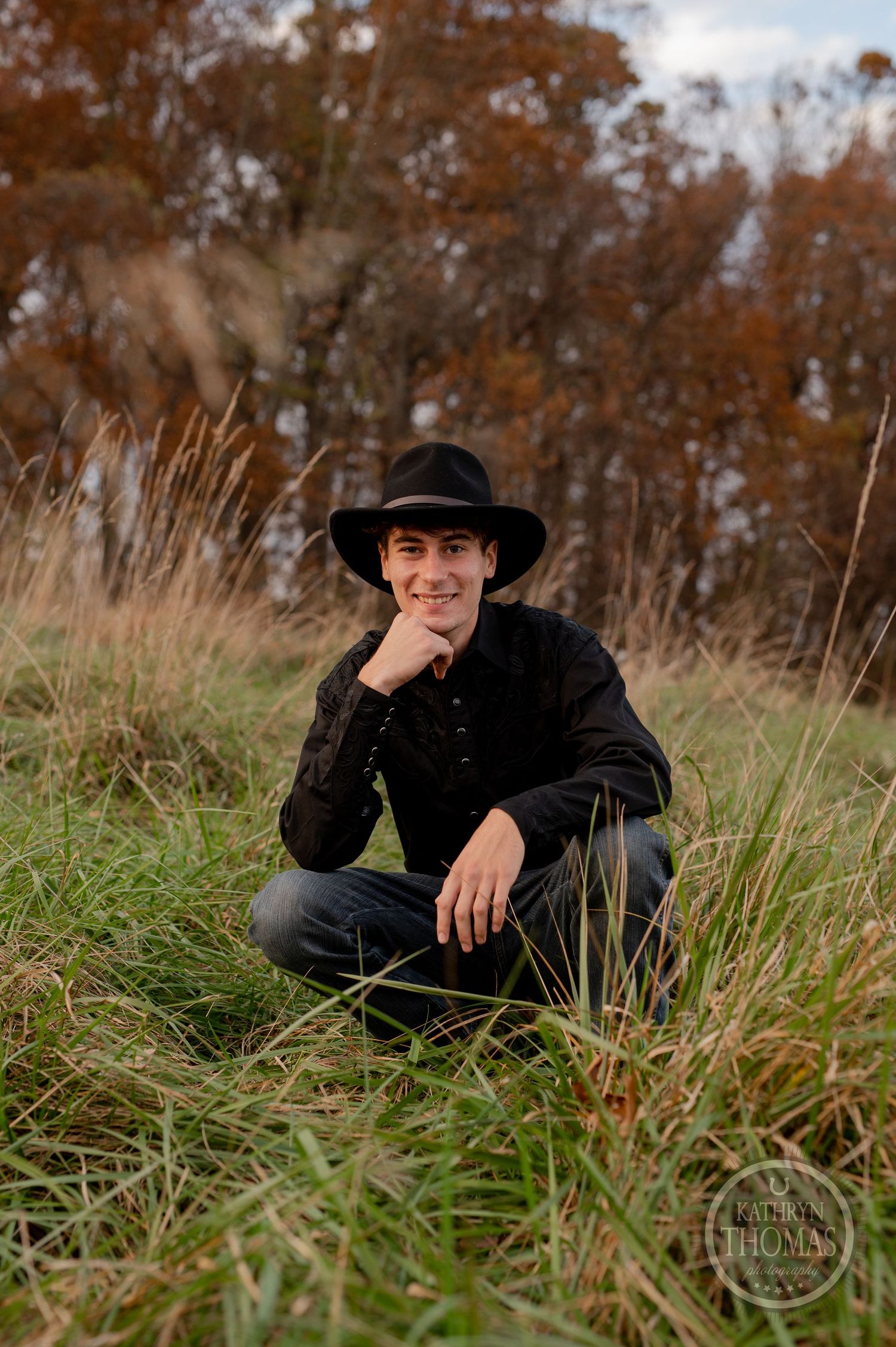 Casual outdoor portrait in western wear sitting among tall autumn grass.