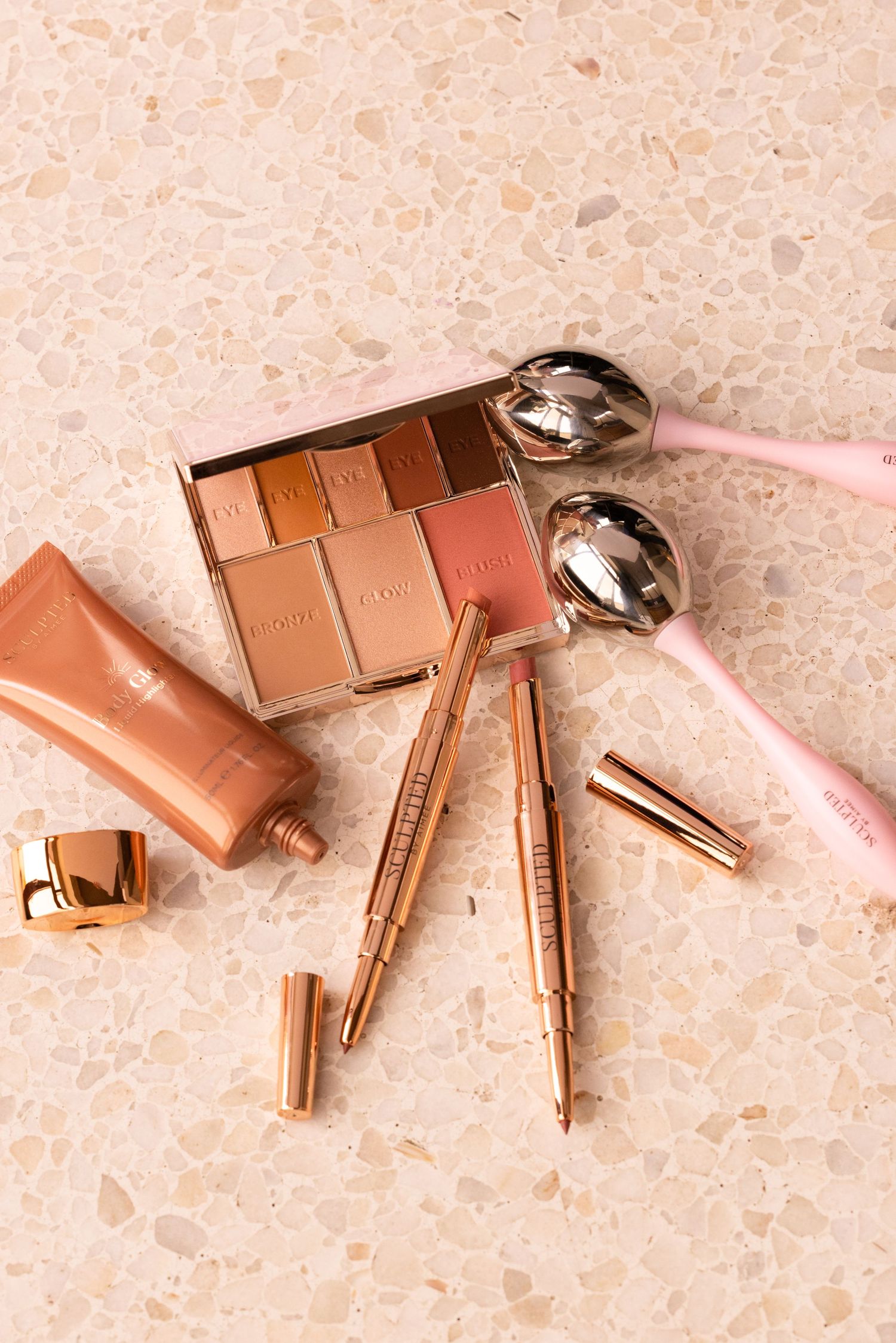 Rose gold makeup collection including eyeshadow palette and brushes on peach terrazzo surface.
