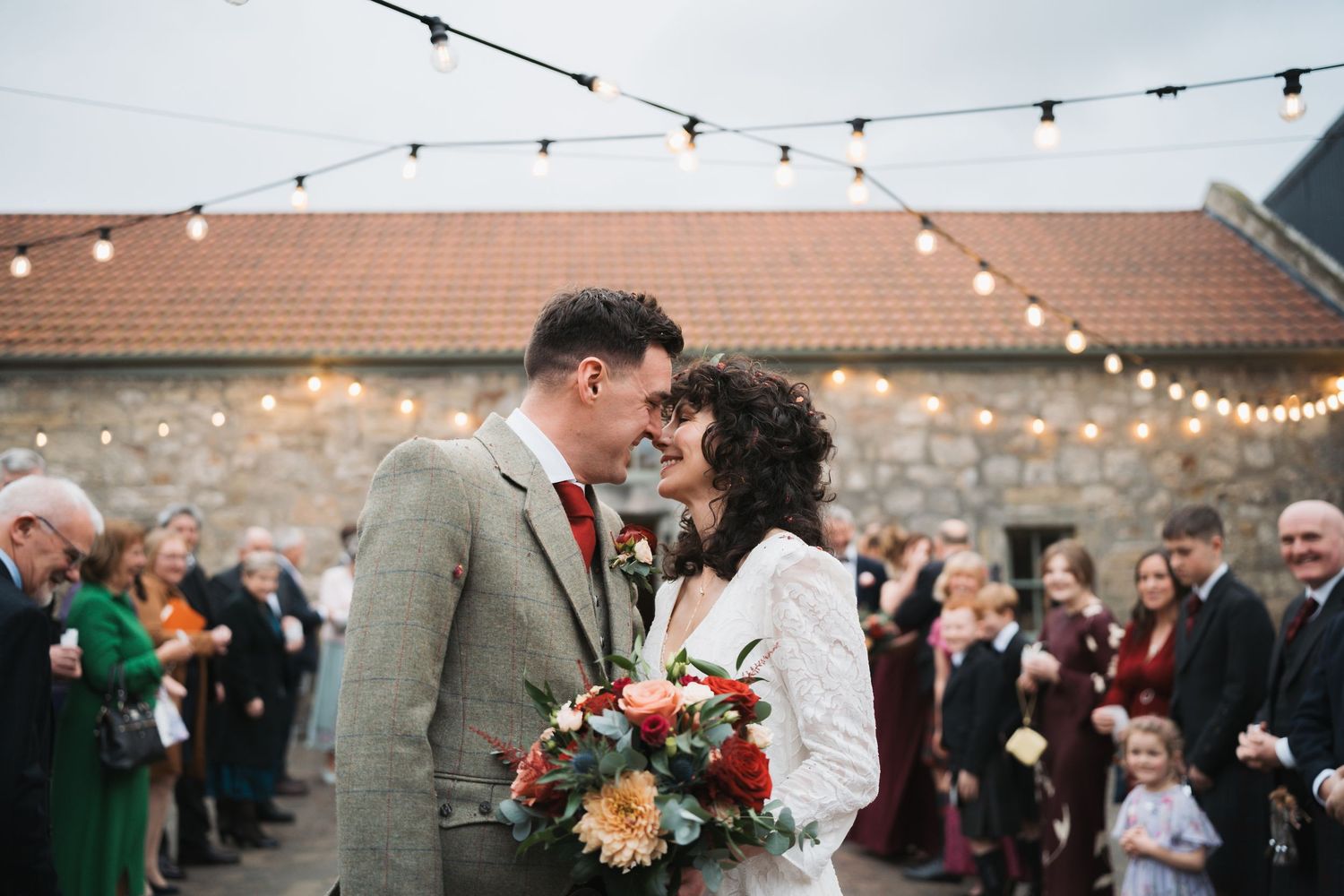 Happy couple shares joyful moment outside stone building decorated with lights for rustic outdoor wedding celebration.