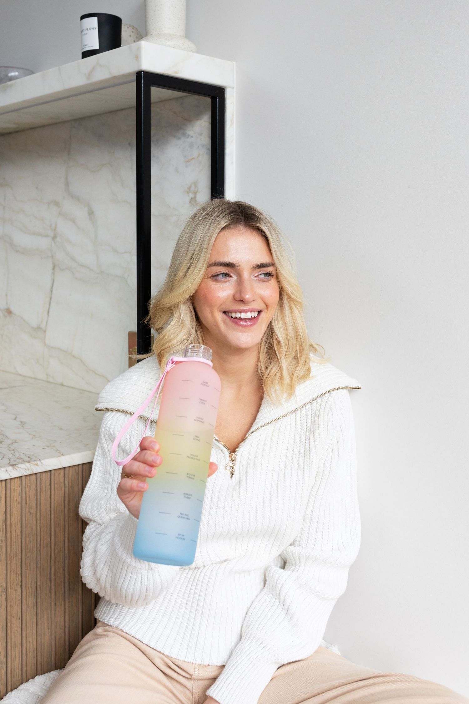 Person in white sweater holding colorful gradient water bottle against white wall.