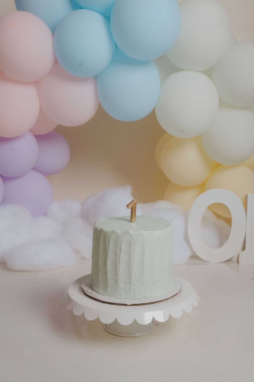 A pastel blue birthday cake with number one candle against pastel balloon backdrop.