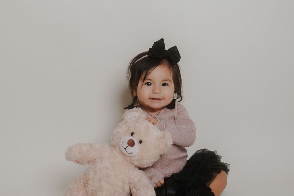 Toddler in pink sweater posing with plush teddy bear against neutral backdrop.