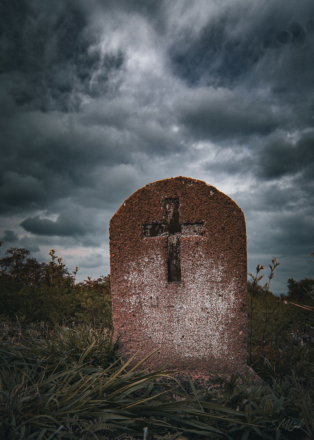 A weathered stone cross marker stands against dramatic stormy clouds in an overgrown field at dusk.
