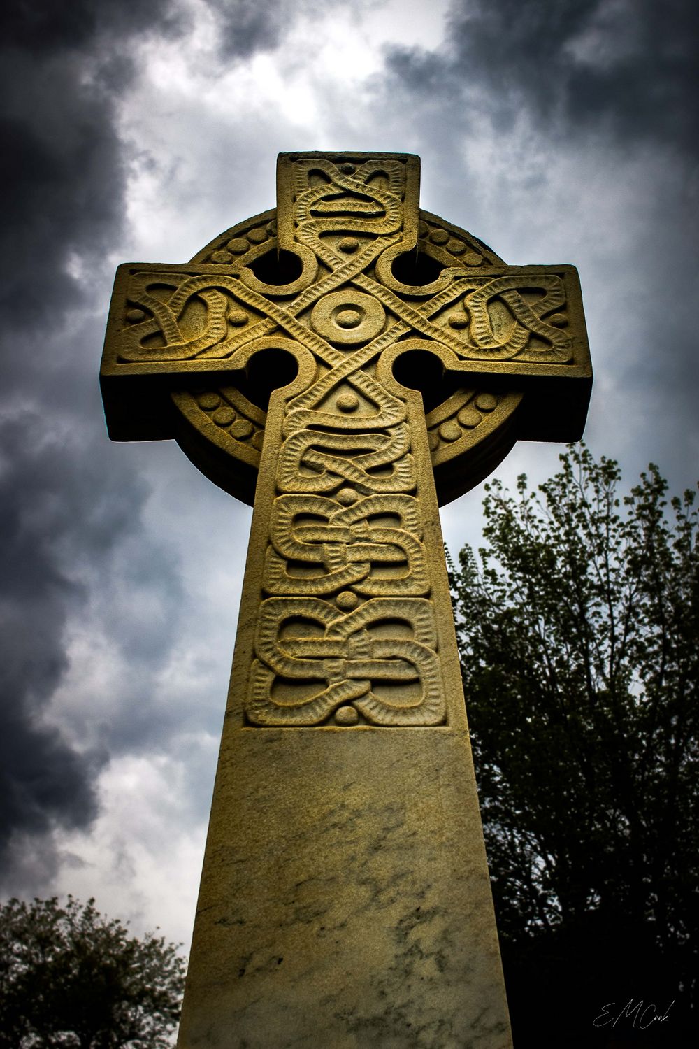 Ancient Celtic stone cross with intricate knotwork patterns stands against dramatic stormy sky with dark clouds.