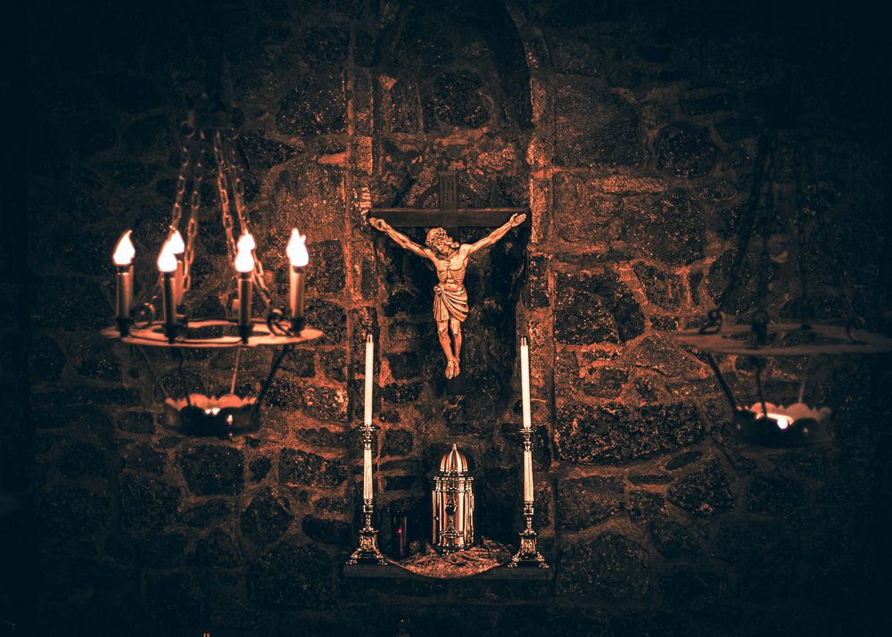 Illuminated candles cast a warm glow on a dark stone wall in a dramatic religious setting.