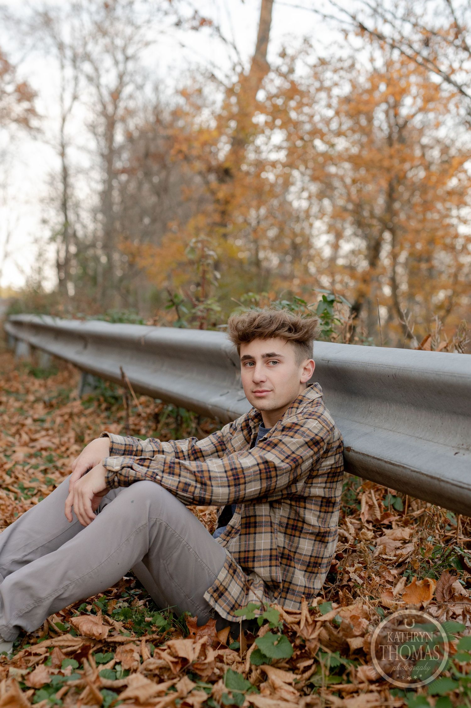 Someone rests against a metal guardrail on a leaf-covered forest path during autumn.