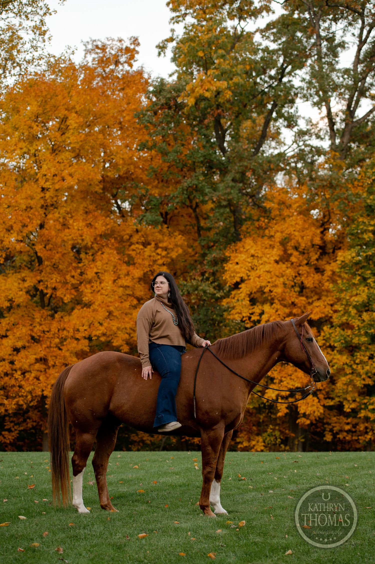 A chestnut horse with white socks stands proudly with a rider against bright orange fall foliage.