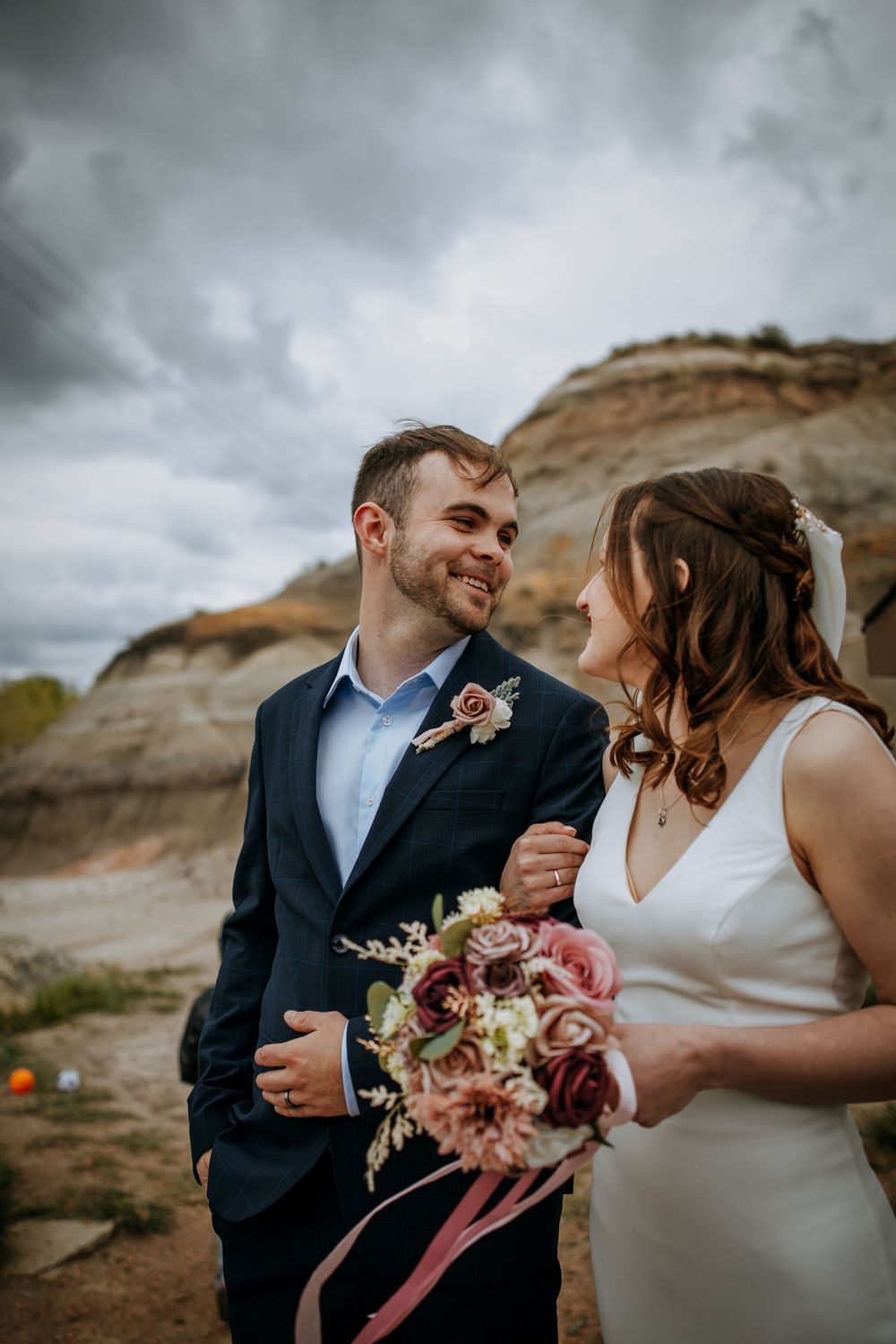 Wedding couple smiles at each other against dramatic rocky landscape with bouquet.