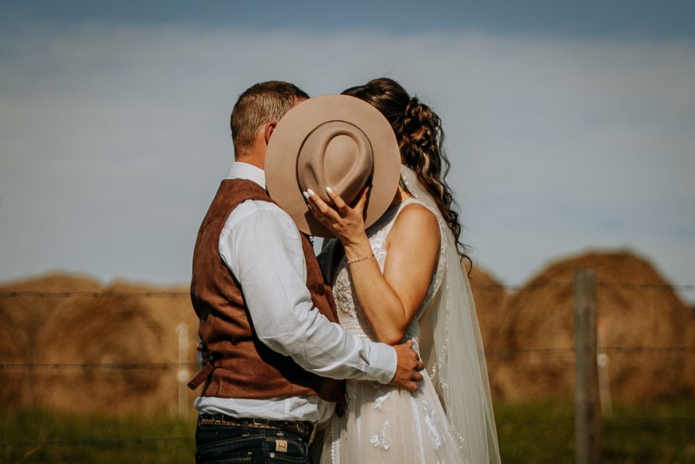 Romantic wedding couple sharing an intimate moment behind a brown cowboy hat in a rustic field.