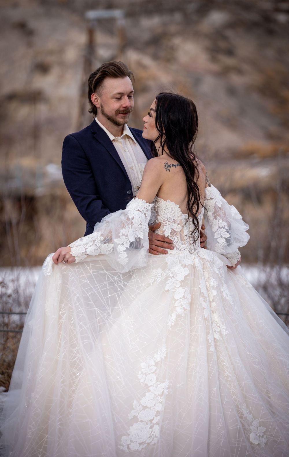 A couple embraces outdoors in winter, with the bride wearing an ornate off-shoulder lace wedding gown.