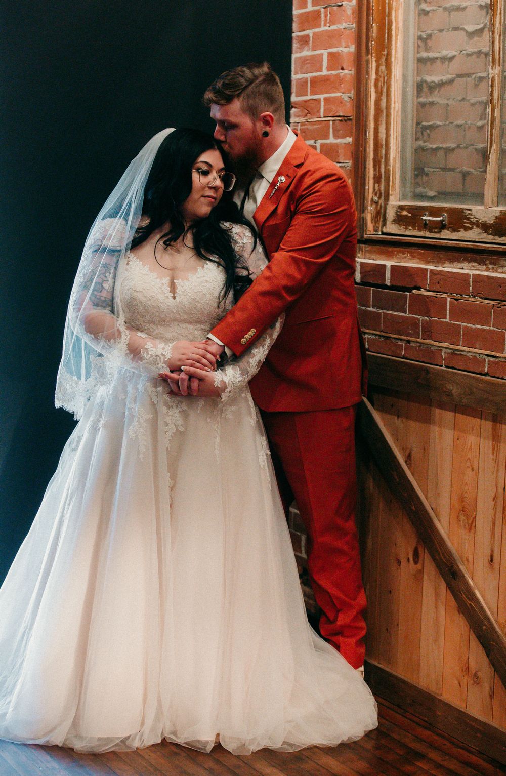 Wedding couple poses against rustic brick wall, bride in white lace gown and groom in red suit.