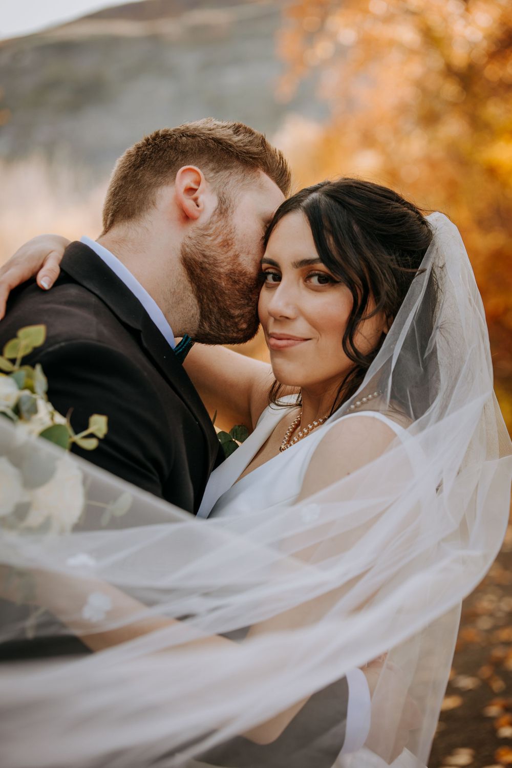Wedding couple embracing outdoors with flowing veil against autumn foliage background.