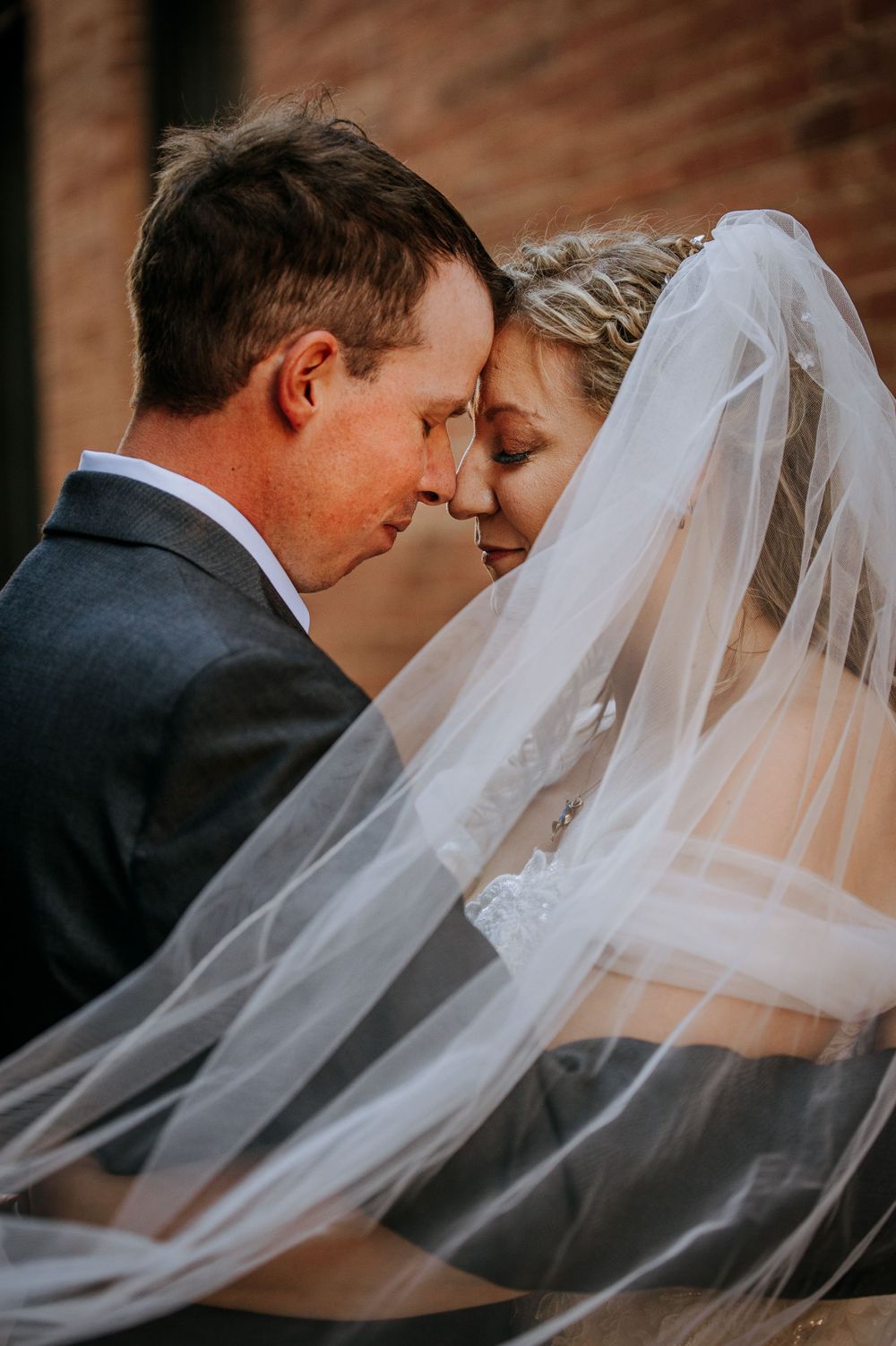 Romantic wedding portrait with couple in an intimate moment as bride's veil flows behind them against brick wall backdrop.