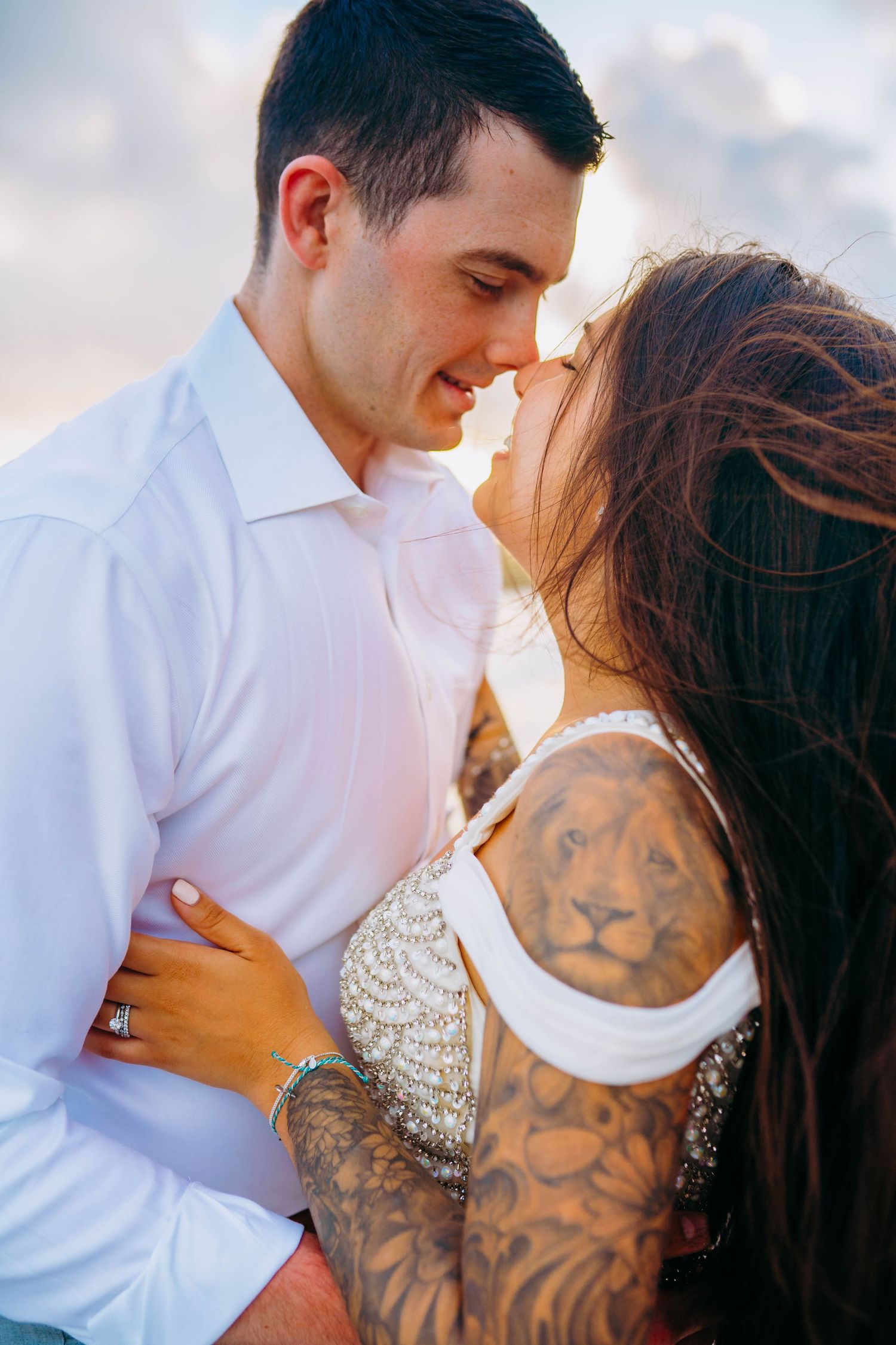 Intimate beach portrait of couple embracing with visible tattoo sleeve and white attire at sunset in Hawaii for beach destination wedding