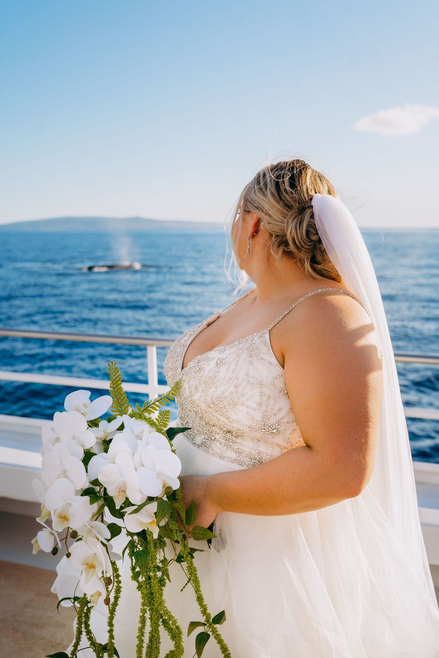 Bride in white dress holds cascading orchid bouquet on boat deck with ocean view and humpaback whale wedding photo on trilogy catamaran