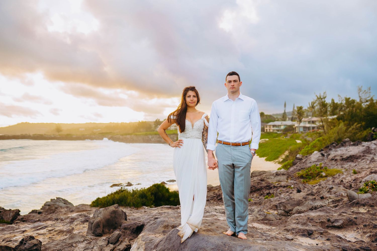 Couple in white attire standing on rocky coastal cliff during sunset in Hawaii for beach destination wedding in maui hawaii.