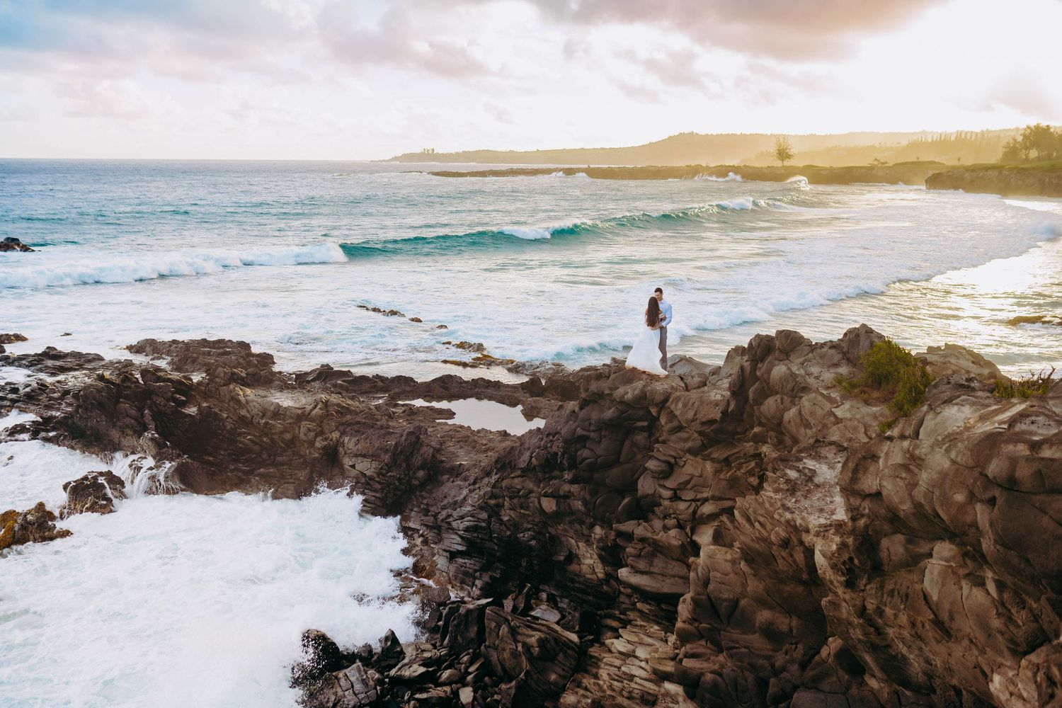 Lone figure stands on dramatic rocky coastline with crashing waves and sunset sky. Stunning wedding photos in maui hawaii on the beach