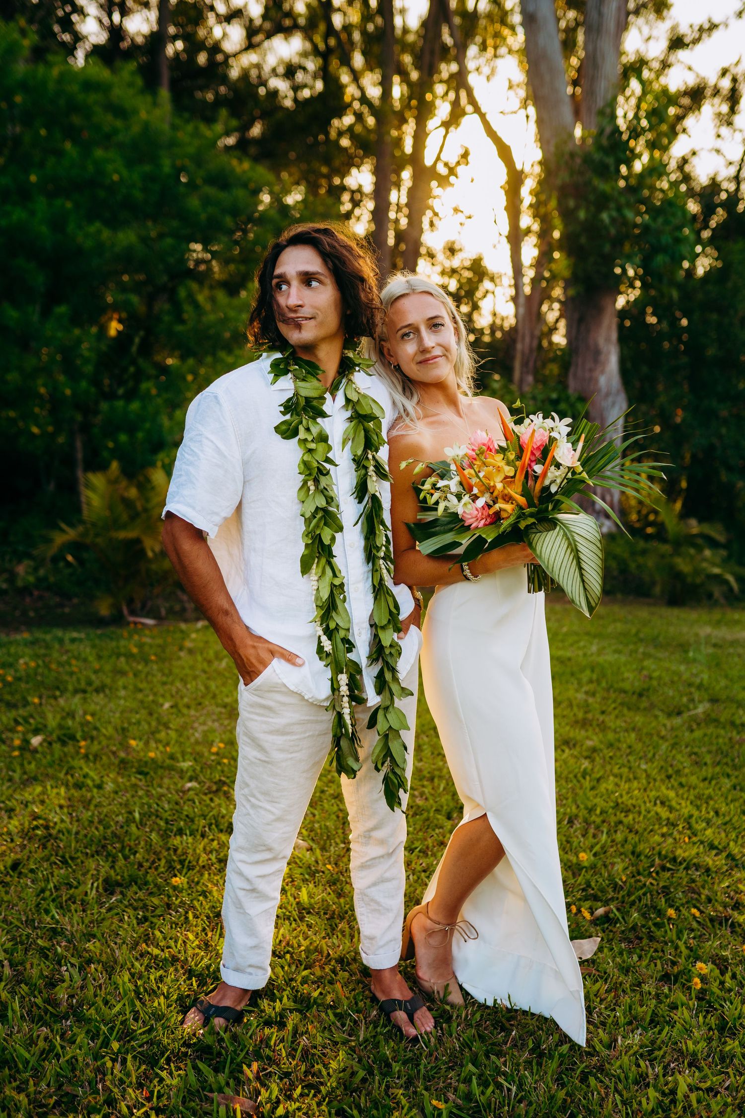 A couple in white wedding attire poses together in a lush garden setting during golden hour sunset. Maui haiku north shore wedding tropical