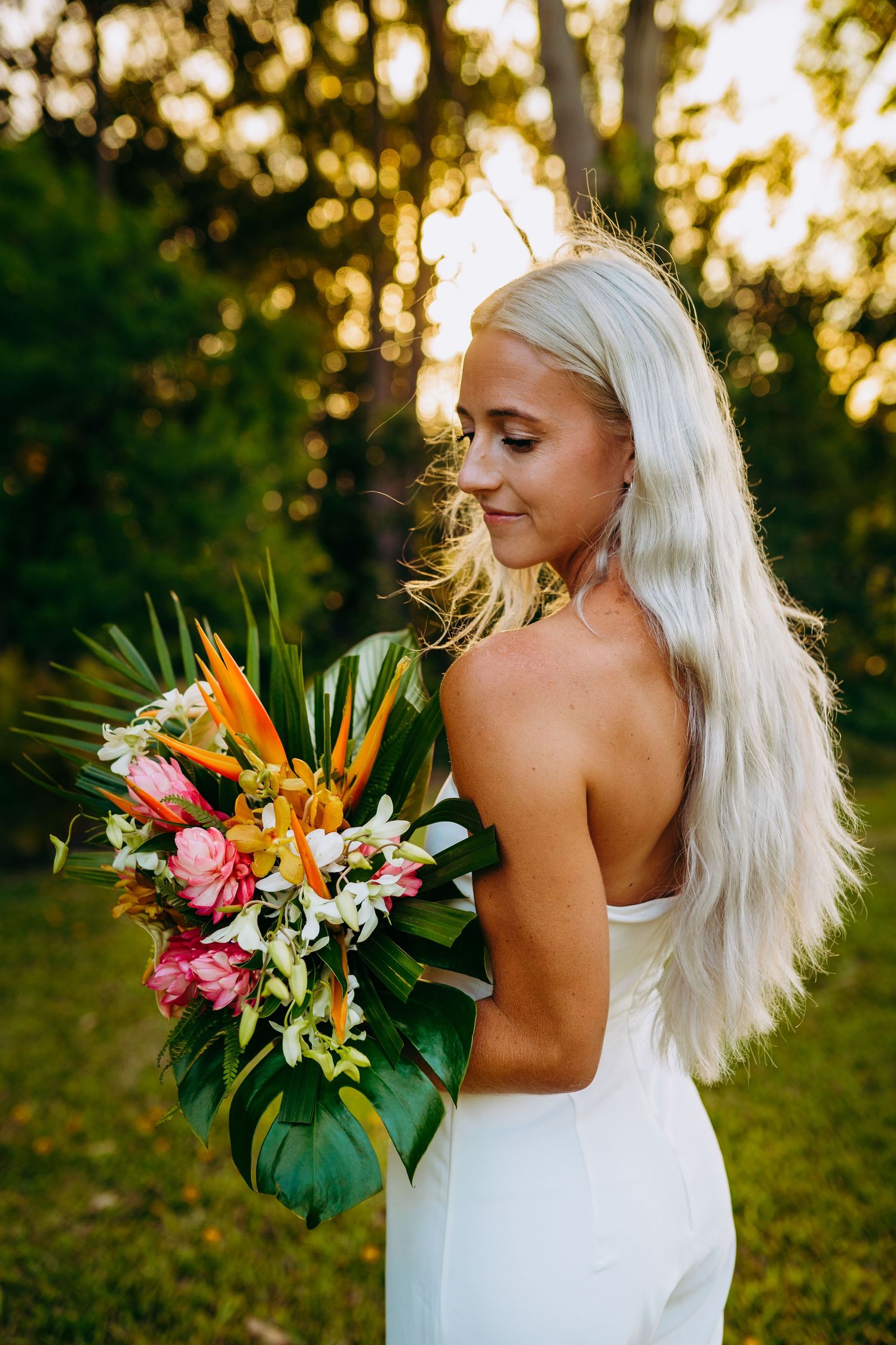 A bride in a white dress poses outdoors at sunset with a vibrant tropical bouquet featuring birds of paradise and lilies. Maui Hawaii Haiku