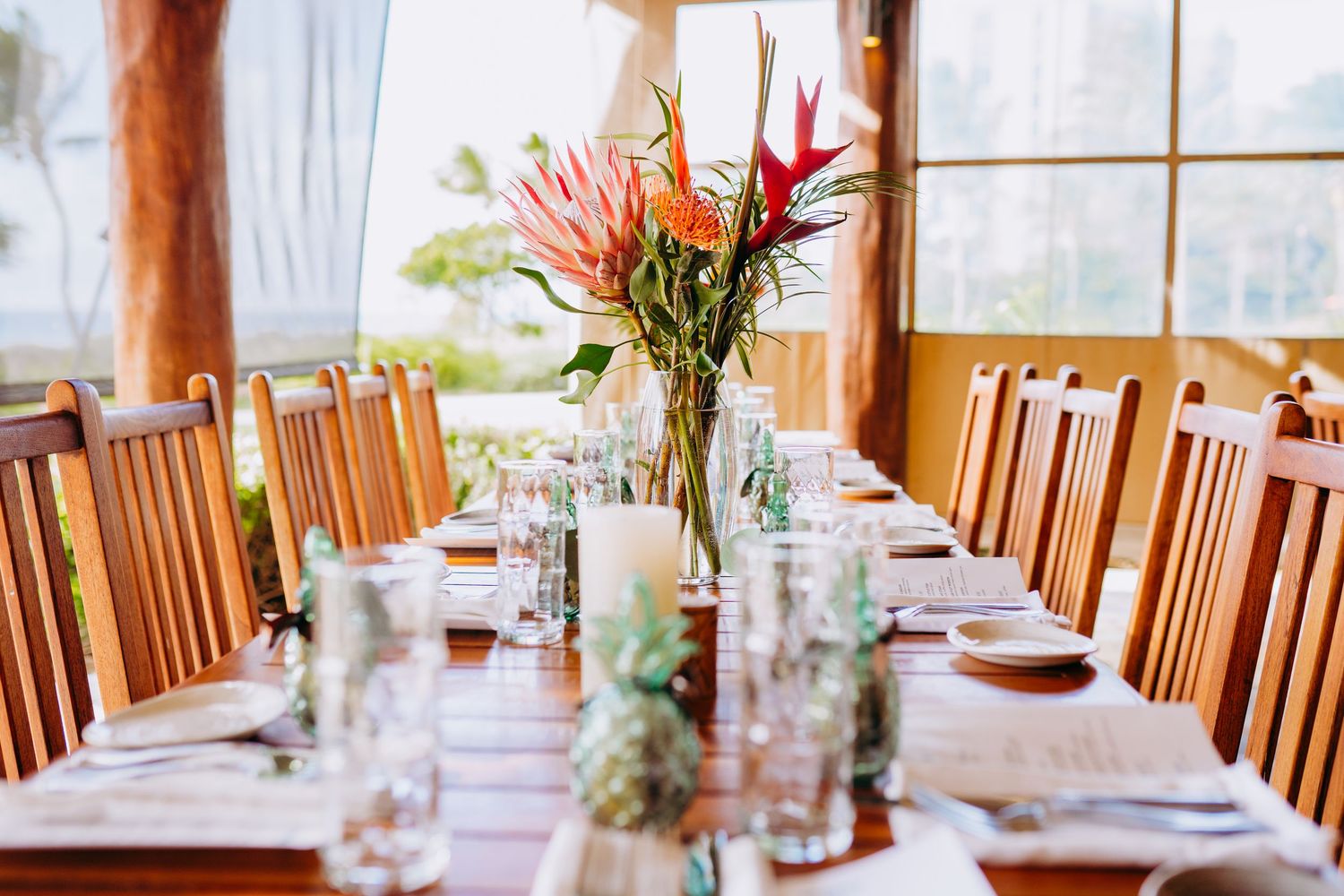 Elegant wedding reception tables set with white linens, wooden chairs and floral centerpieces in a bright sunlit room. Duke's Maui wedding