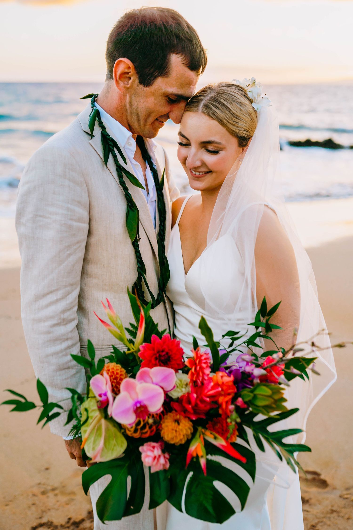 Romantic beach wedding couple sharing an intimate moment with tropical flowers and monstera leaves at sunset in Hawaii for beach destination