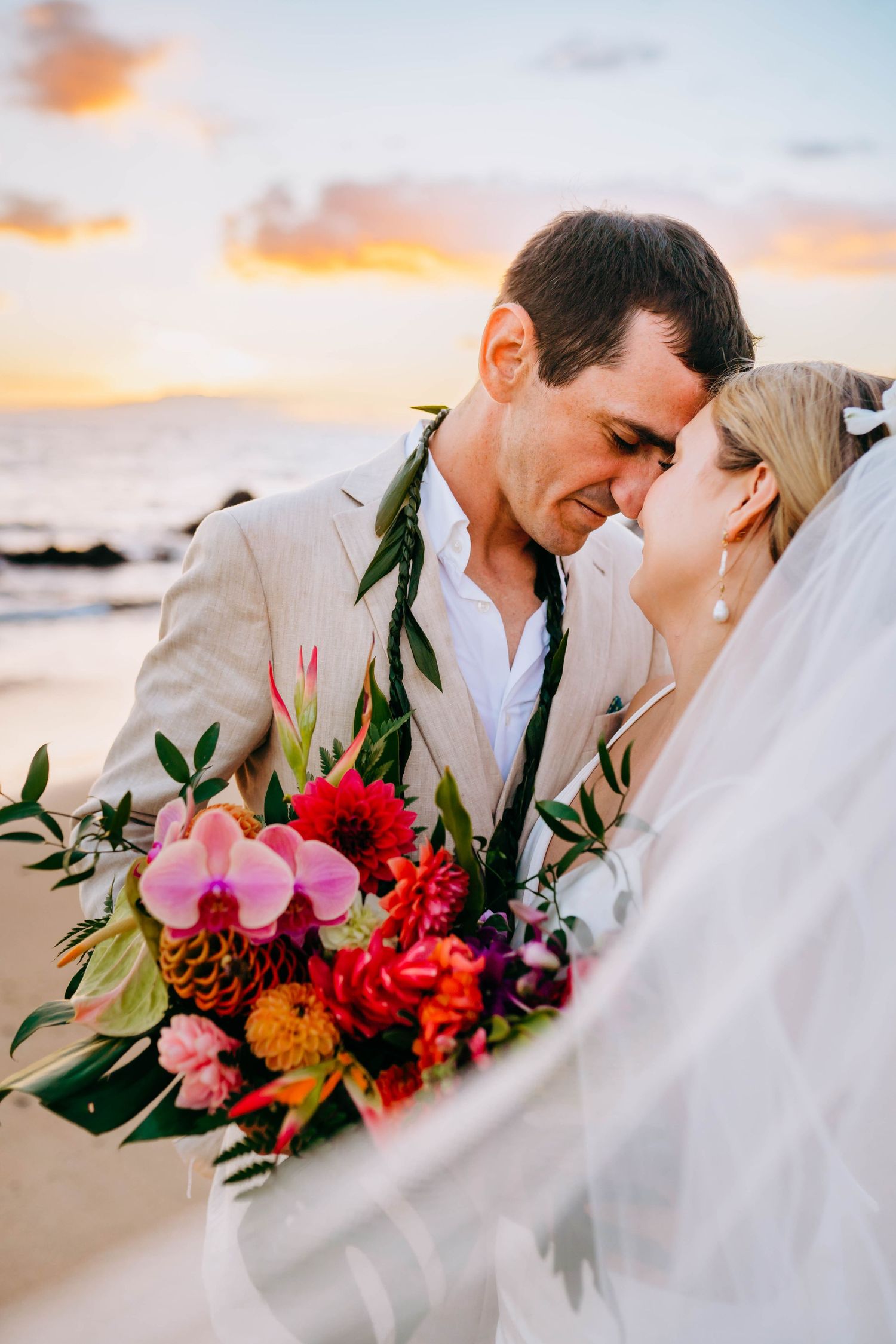 Newlyweds share tender moment on beach with vibrant tropical bouquet during golden hour in Hawaii for beach destination wedding in maui