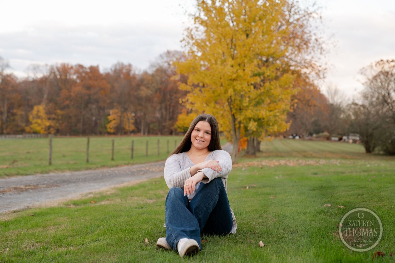 A relaxed outdoor pose on grass beside a country path with autumn trees in the background.