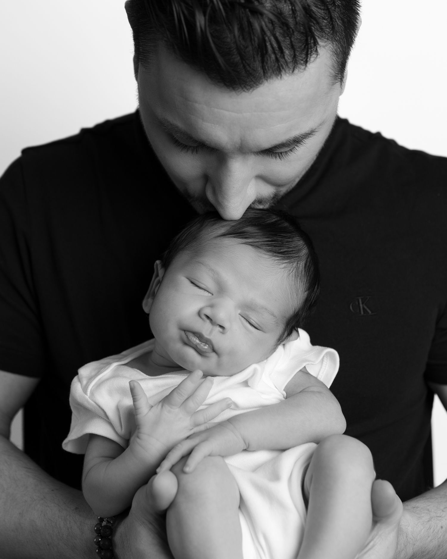 Black and white portrait showing tender moment between a father holding and kissing his sleeping baby.