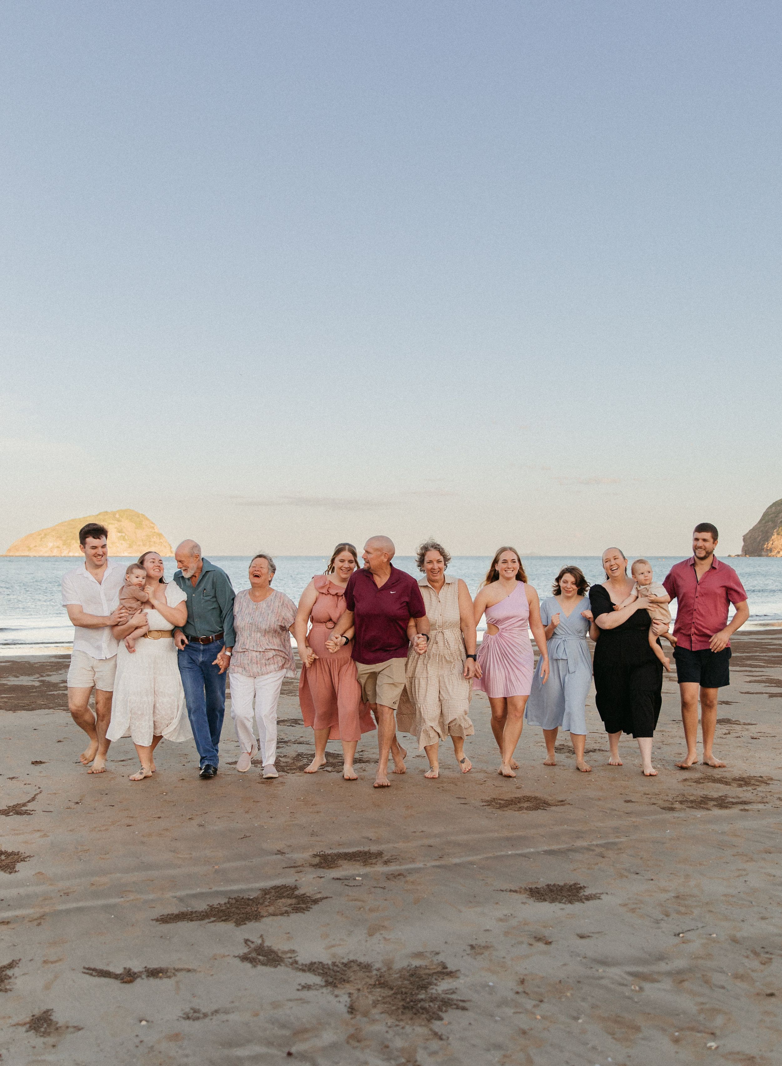Large family group smiling and walking together on a sandy beach at sunset with ocean waves and rocky headland behind.