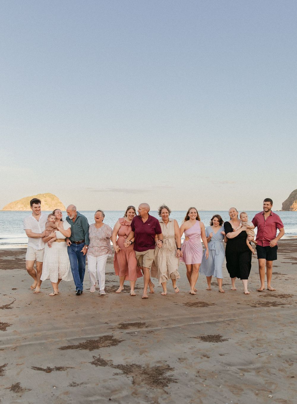 Large family group smiling and walking together on a sandy beach at sunset with ocean waves and rocky headland behind.