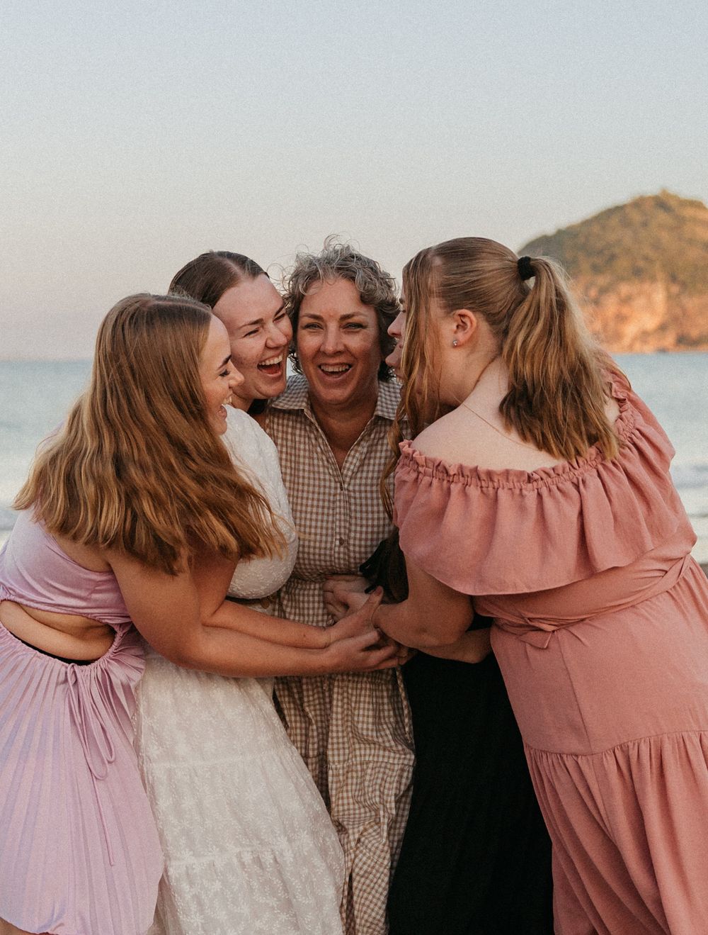 A joyful group of women hugging and laughing together on a beach at sunset.