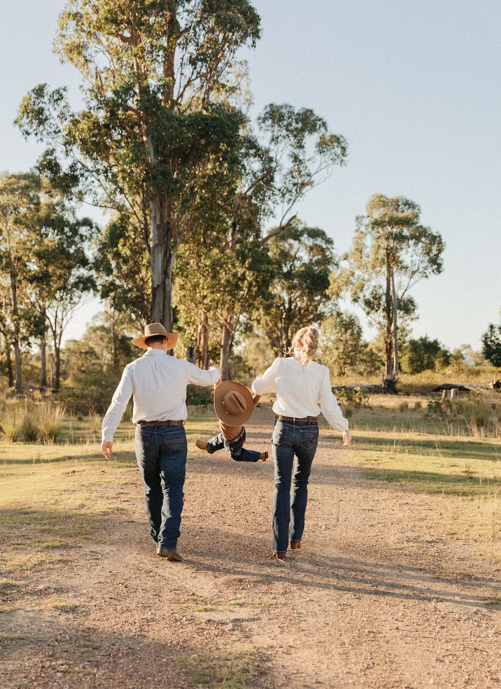 Family of three walking down a dirt path surrounded by eucalyptus trees, parents swinging child between them.