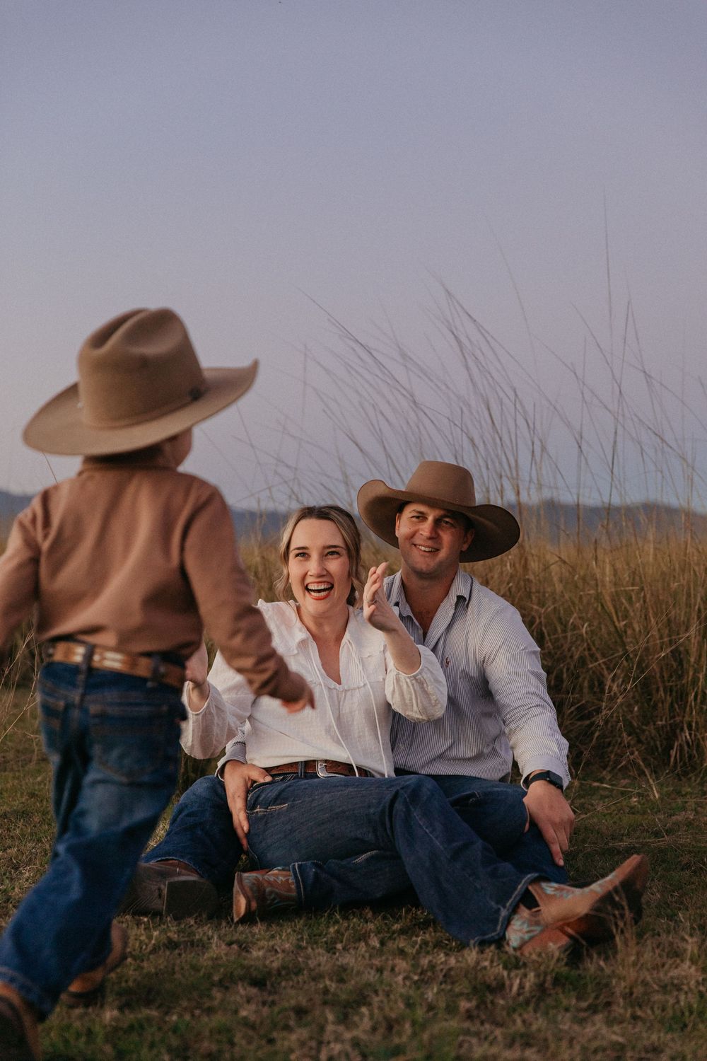 Happy couple in cowboy hats sitting in grass laughing as young child runs toward them outdoors.