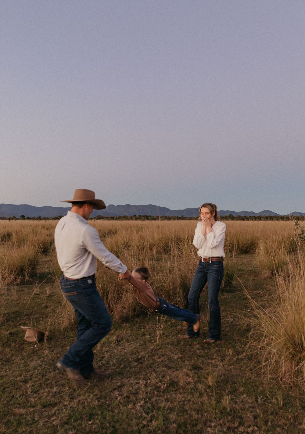 Father in cowboy hat swinging child by arms while mother laughs in open golden grassland field.