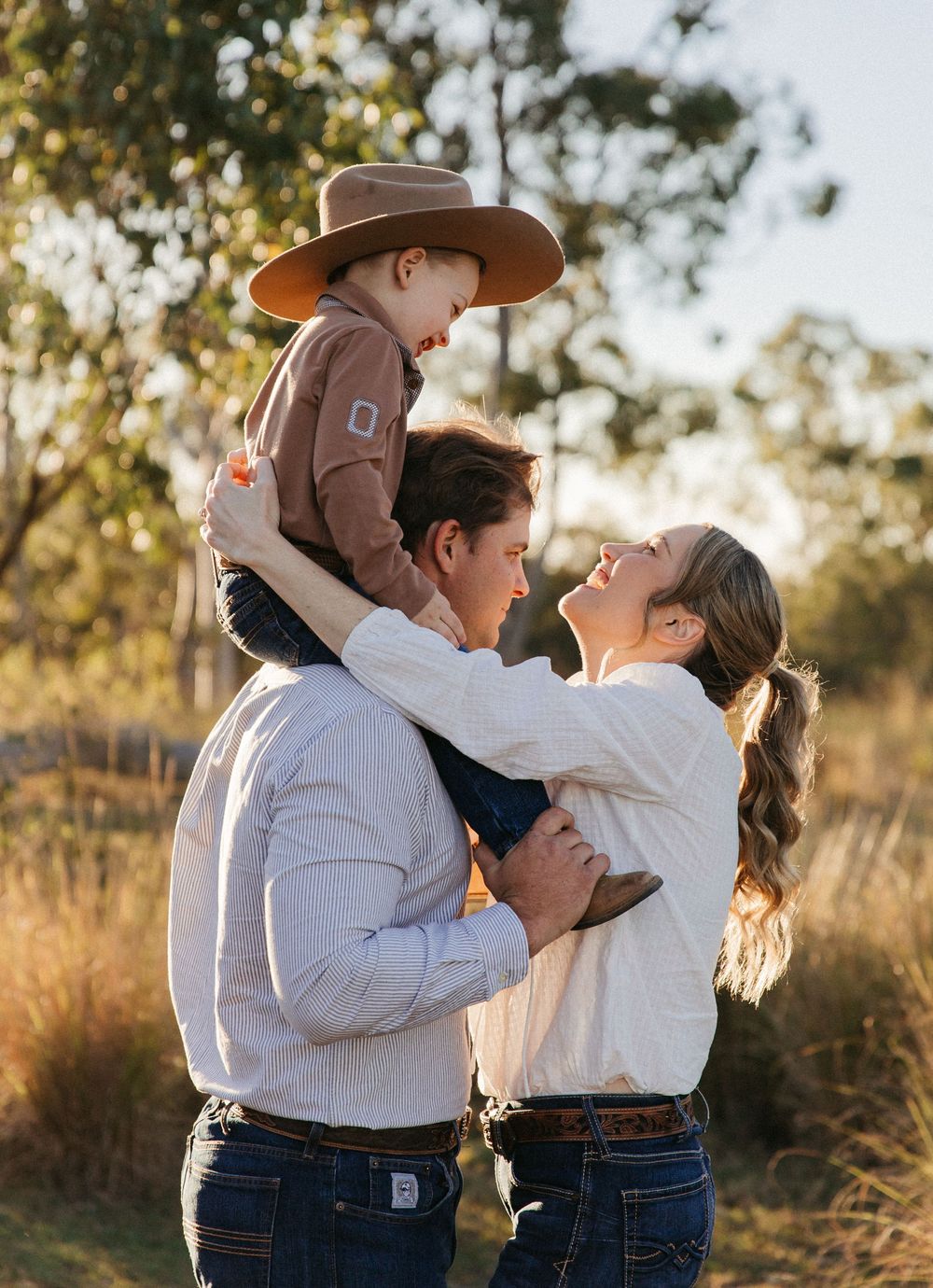 A young family shares a joyful moment outdoors, with parents lifting their toddler in a cowboy hat at golden hour.