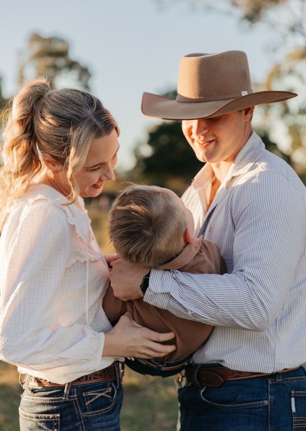 A young couple in jeans and white shirts lovingly hold their toddler outdoors in golden hour light.