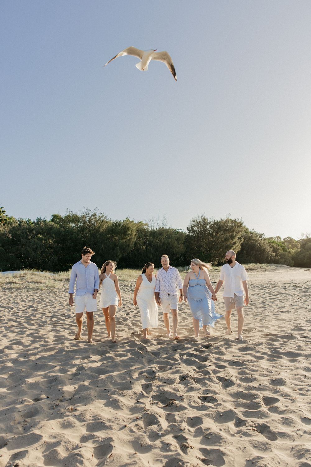 Group of six people in white and blue outfits walking on sandy beach with a seagull flying overhead.