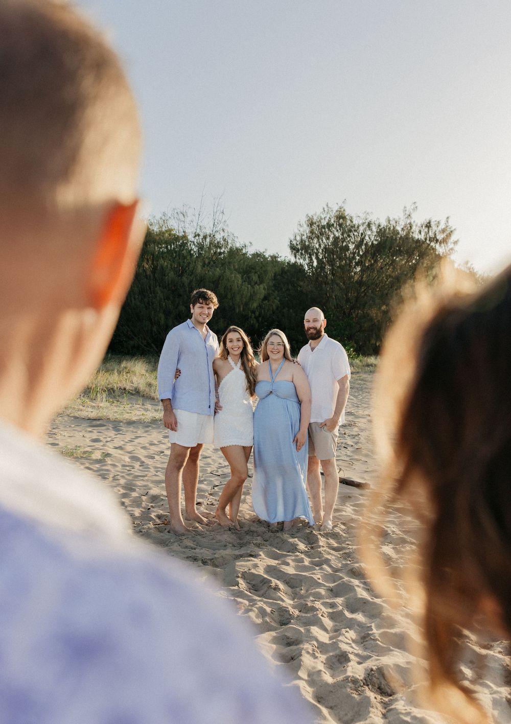 Four people pose on sandy beach at golden hour, wearing light blue and white outfits, with green trees in background.