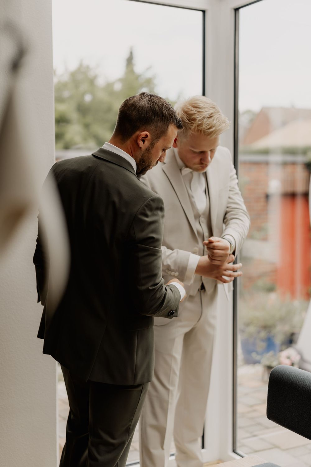 Two people in contrasting suits helping with wedding day preparations near a window.