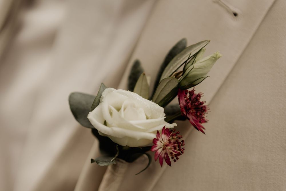 A delicate white rose and burgundy floral boutonniere with eucalyptus leaves against a cream background.
