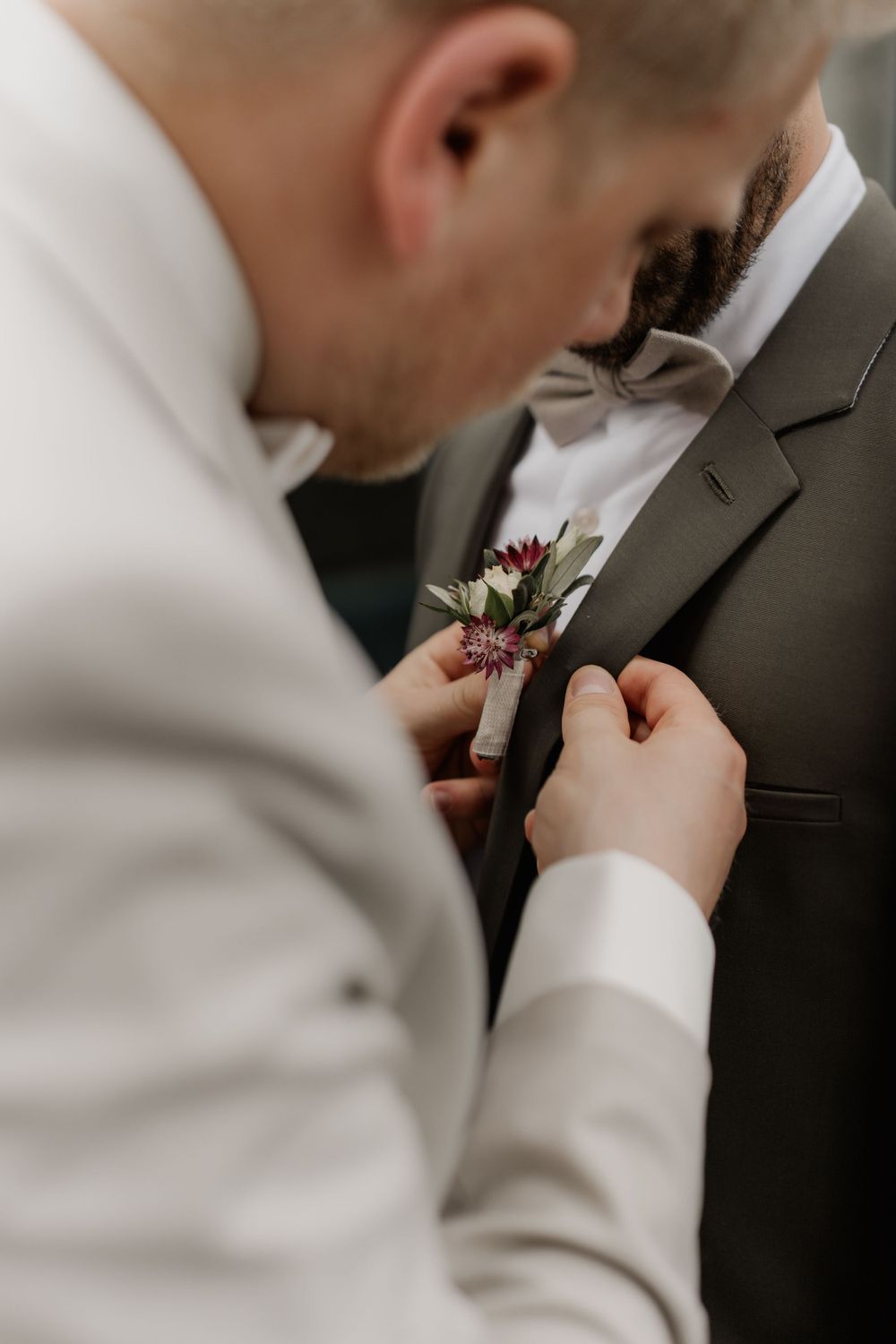 Close-up shot of hands pinning a boutonniere with white and burgundy flowers onto a dark suit.