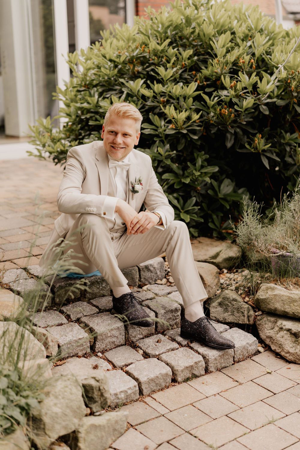 A person in a light beige suit sitting on stone steps next to a green bush.
