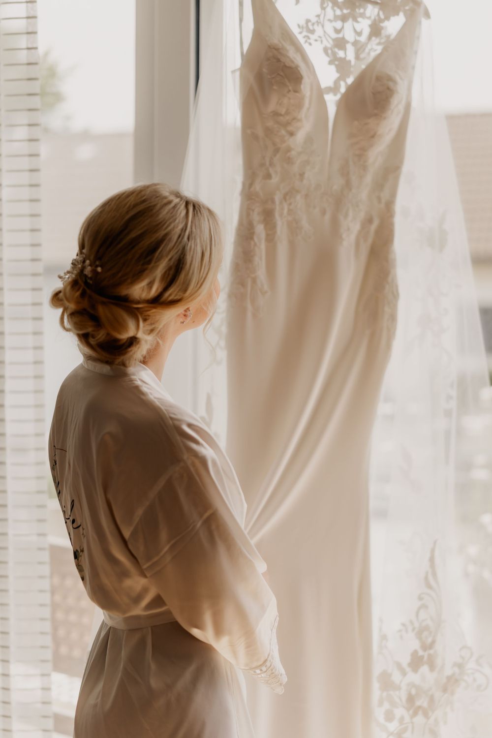 Silhouette of someone in a robe looking at an elegant white wedding dress hanging by a window.