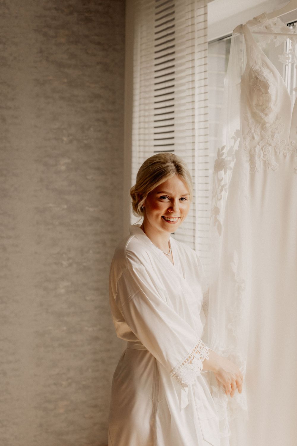 Someone wearing a white robe standing by a window near an elegant wedding dress.