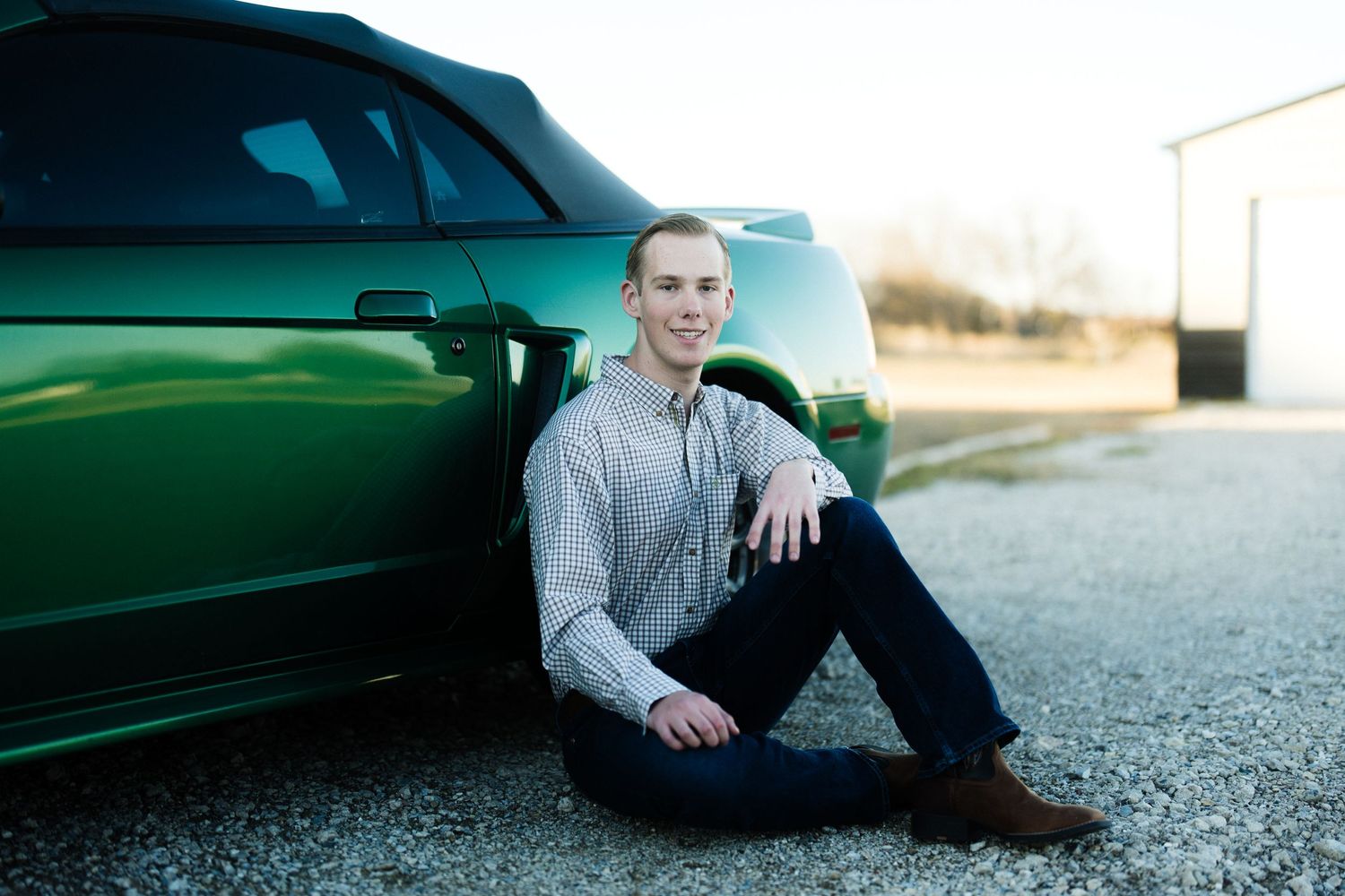 Person in grey sweater sitting beside green car on pavement at sunset.