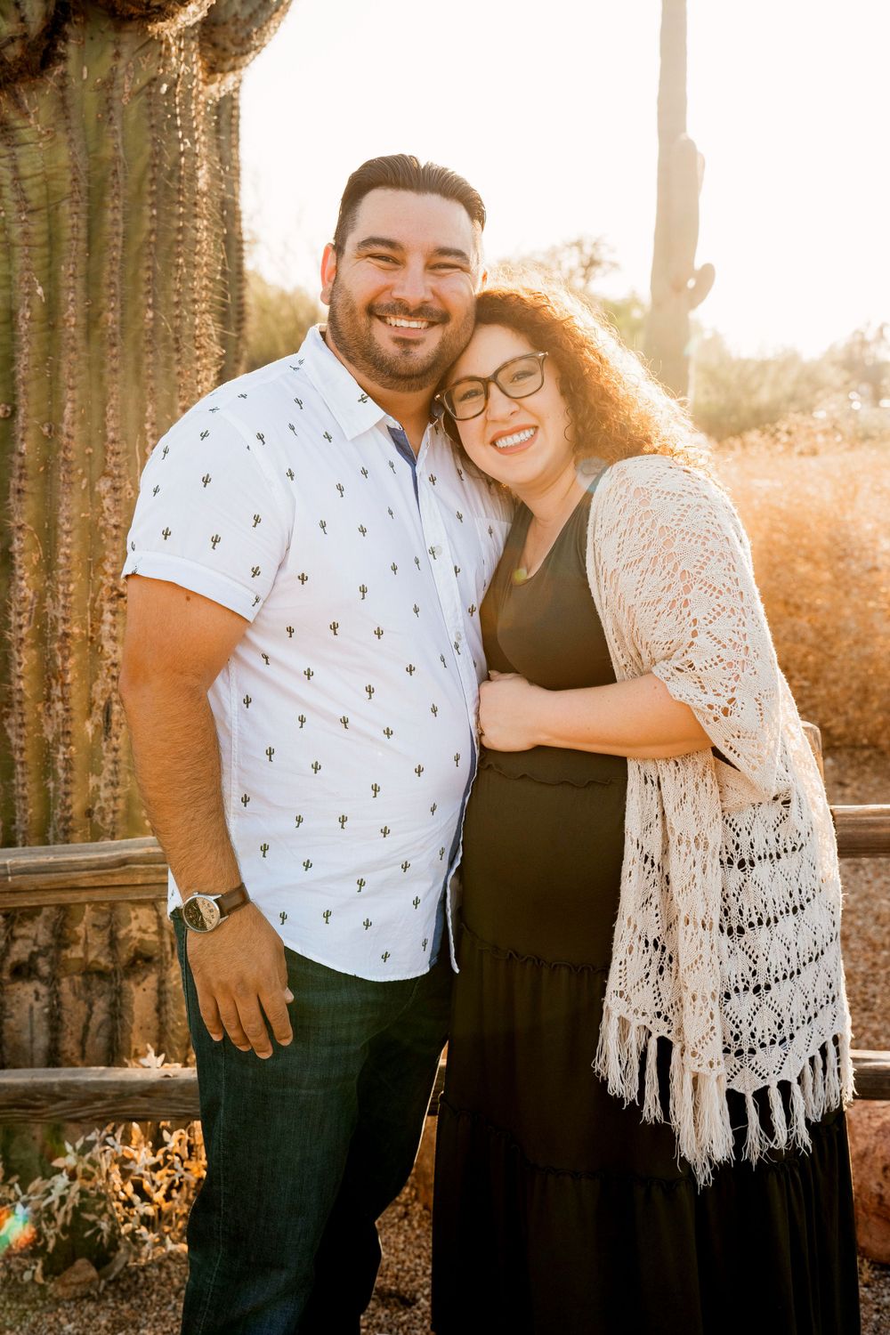 Expectant couple embracing and smiling in front of desert cactus during golden hour.