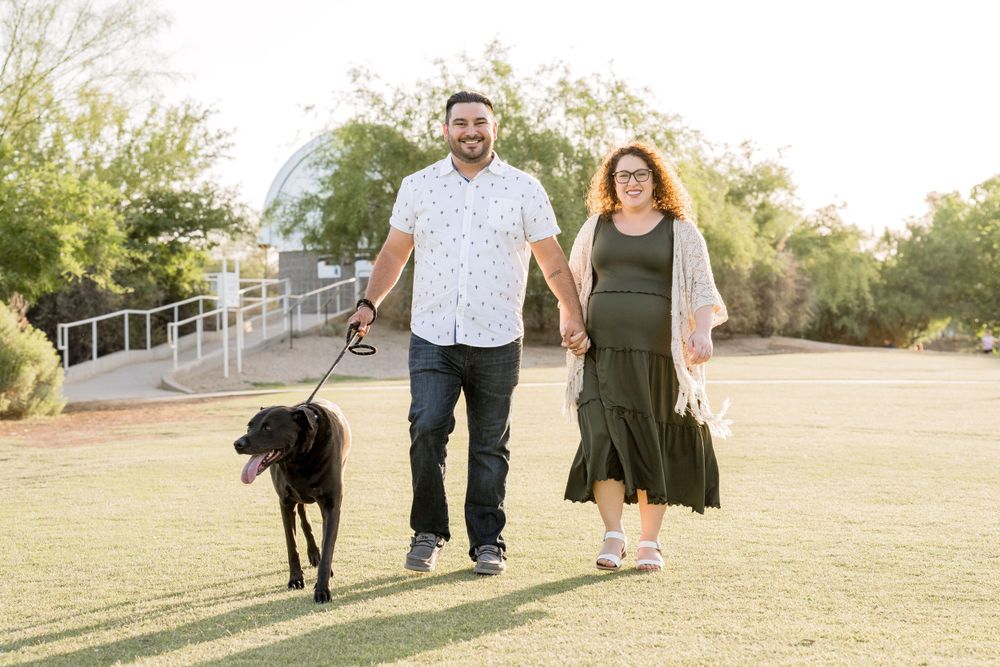 Couple walking their black dog on grass path with desert landscape in background.