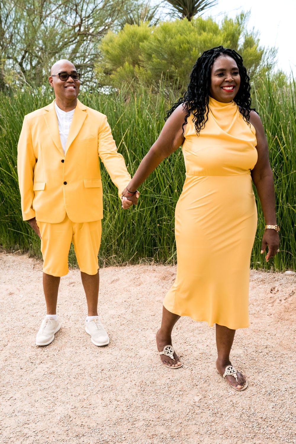 A couple in matching yellow outfits holding hands on a path with greenery in the background.