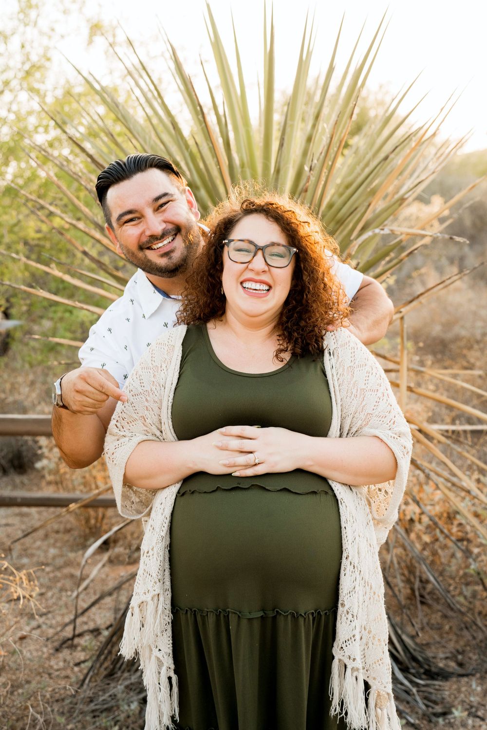 Joyful maternity photo of couple laughing together with desert plants in background.